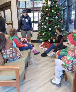 A person in uniform speaks to a group of children sitting on benches near a decorated Christmas tree indoors.