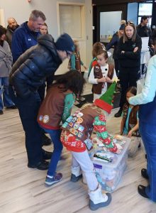 A group of children in uniforms and an elf costume gather around a box in an indoor setting, with adults standing nearby.