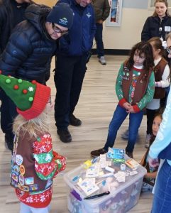 Children in uniform gather around a clear bin filled with various items, as several adults in coats stand nearby in a room with wooden flooring.