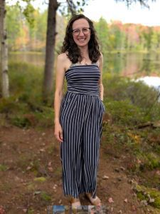 A person standing outdoors near a lake, wearing a striped jumpsuit and glasses, smiles at the camera. Trees and foliage are in the background.