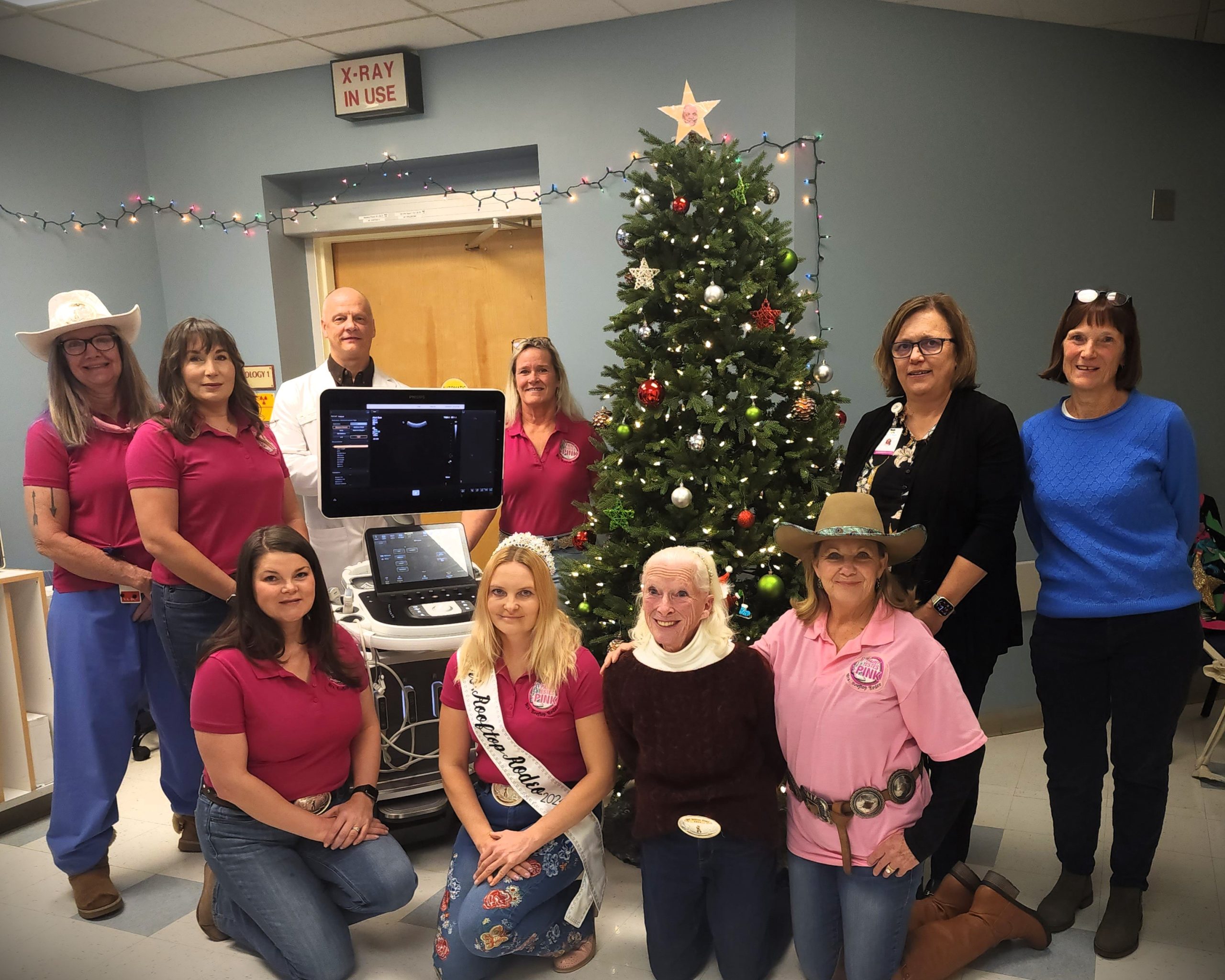 Group of people posing in front of a Christmas tree and medical equipment, some wearing cowboy hats and pink shirts, in a decorated room.