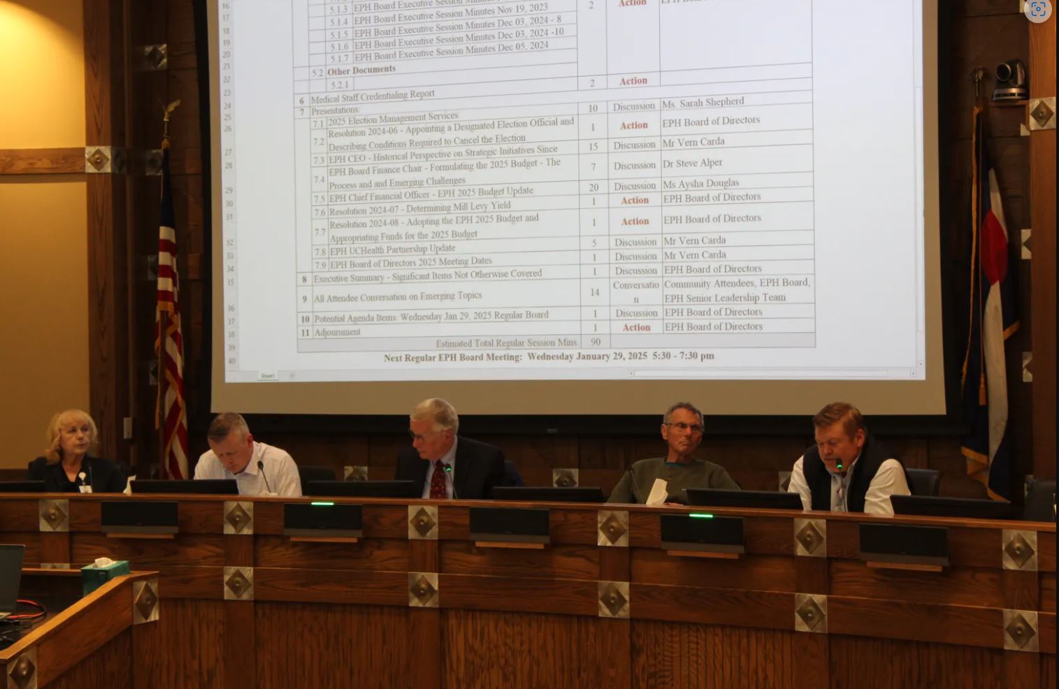 Five people sit at a conference table with a large document projected on a screen behind them in a wood-paneled room.