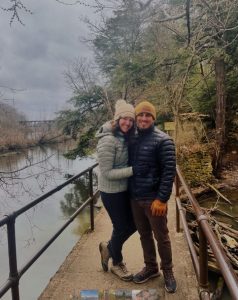 A couple stands on a bridge over a calm river, bundled in winter jackets and hats with trees and an overcast sky in the background.