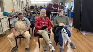 Three seated older men in casual clothing face forward; a woman is seated behind them. In the background, several people are working in a spacious room with wooden floors.