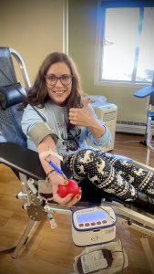 A woman donates blood, holding a red stress ball and giving a thumbs-up, while sitting on a reclined chair.