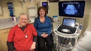 Two women, one in a red top and the other in a blue top, sit beside an ultrasound machine in a medical room.