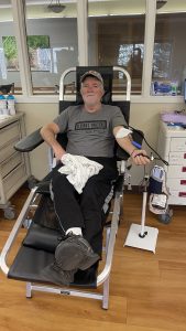 A man sits in a chair donating blood, with a needle in his arm and a blood bag beside him, inside a medical facility.