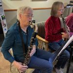 Two women seated and playing clarinets, focused on reading sheet music. One wears a green shirt, the other a red velvet top.