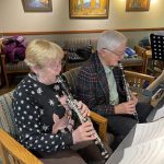 Two elderly people seated indoors, playing clarinets with sheet music on stands in front of them. Paintings and coats are visible in the background.