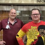 Two men stand indoors. One wears a red polo shirt with an ID badge, and the other wears glasses and a colorful sweater with "Say Merry Xmas Again" written on it, featuring a fictional character.