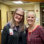 Two women smiling in a lobby area; one wears glasses and a dark cardigan, the other a red plaid shirt.