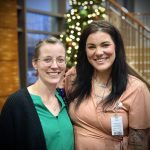 Two people smiling, standing side by side in a brightly lit indoor setting. A decorated Christmas tree and staircase are in the background.