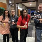 Four people are standing in a lobby area, smiling at the camera. Three are wearing work badges and casual clothes.