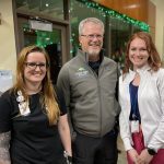 Three people stand indoors, smiling at the camera. The man in the middle wears glasses and a jacket, while the two women on either side wear casual clothing.