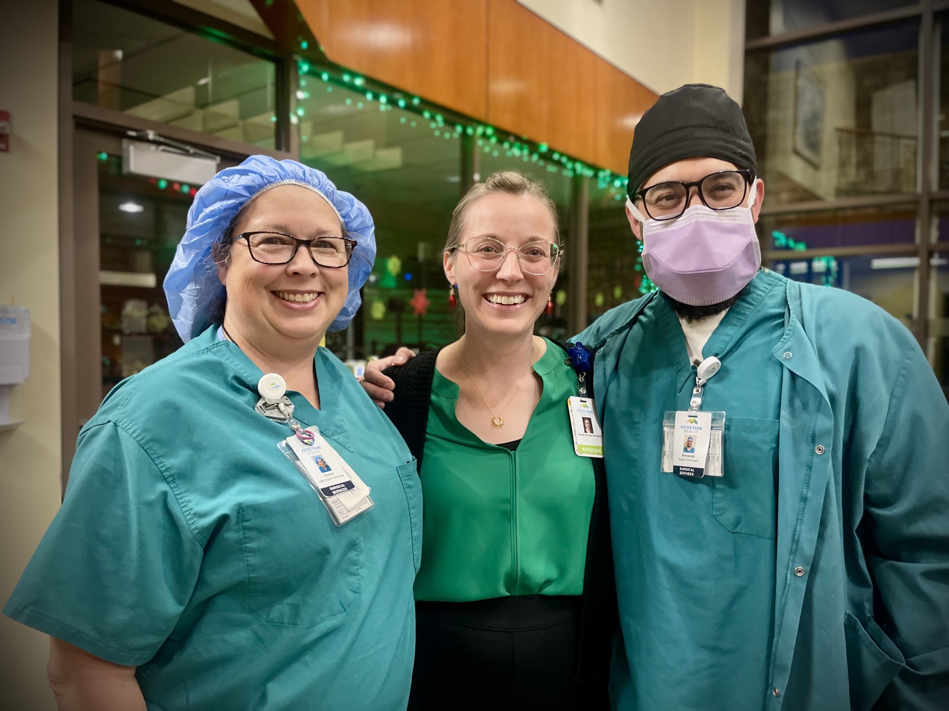 Three healthcare workers smile while posing together indoors. Two are wearing scrubs, and one is wearing a green top with a blazer.