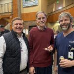 Three men smiling, standing together indoors at an event. One man is holding a slice of cake, and another is holding a can. People are in the background.