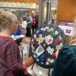 A woman spins a holiday-themed prize wheel decorated with festive symbols at an indoor event. Others watch in the background.