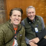 Two men sitting and smiling, both wearing jackets. One man has a sling on his arm. They are in an indoor setting with wooden and stone wall panels.