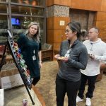 Three people stand around a board with colorful pins in an office lobby, observing or discussing them.