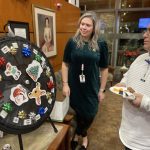 Two people stand by a spinning prize wheel adorned with holiday decorations. One person watches as the other holds a plate with food.