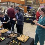 People gather around a table with assorted snacks and desserts at a social event. Some wear healthcare uniforms.