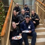 A group of seven people, sitting on stairs and holding plates of food, poses for a photo indoors near a decorated Christmas tree.