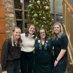 Four women stand smiling in front of a decorated Christmas tree indoors.