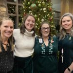 Four women stand in front of a decorated Christmas tree, smiling at the camera. They are indoors, wearing casual attire.