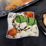 A bowl of mozzarella balls, cherry tomatoes, and basil leaves drizzled with balsamic glaze, served on a black tablecloth next to a tray of cookies and a platter of sliced fruit.