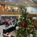 A group of people socializing near a decorated Christmas tree in a two-story lobby with a buffet table set up.