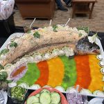 A platter with a large, cooked salmon surrounded by colorful vegetable garnishes, sliced cucumber, onions, and peppers, displayed on a buffet table.