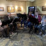 A group of six people seated, playing clarinets and saxophones in a cozy room with framed art on the walls.