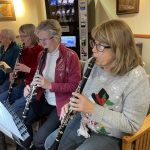 A group of four women sits and plays clarinets indoors, with a vending machine in the background.