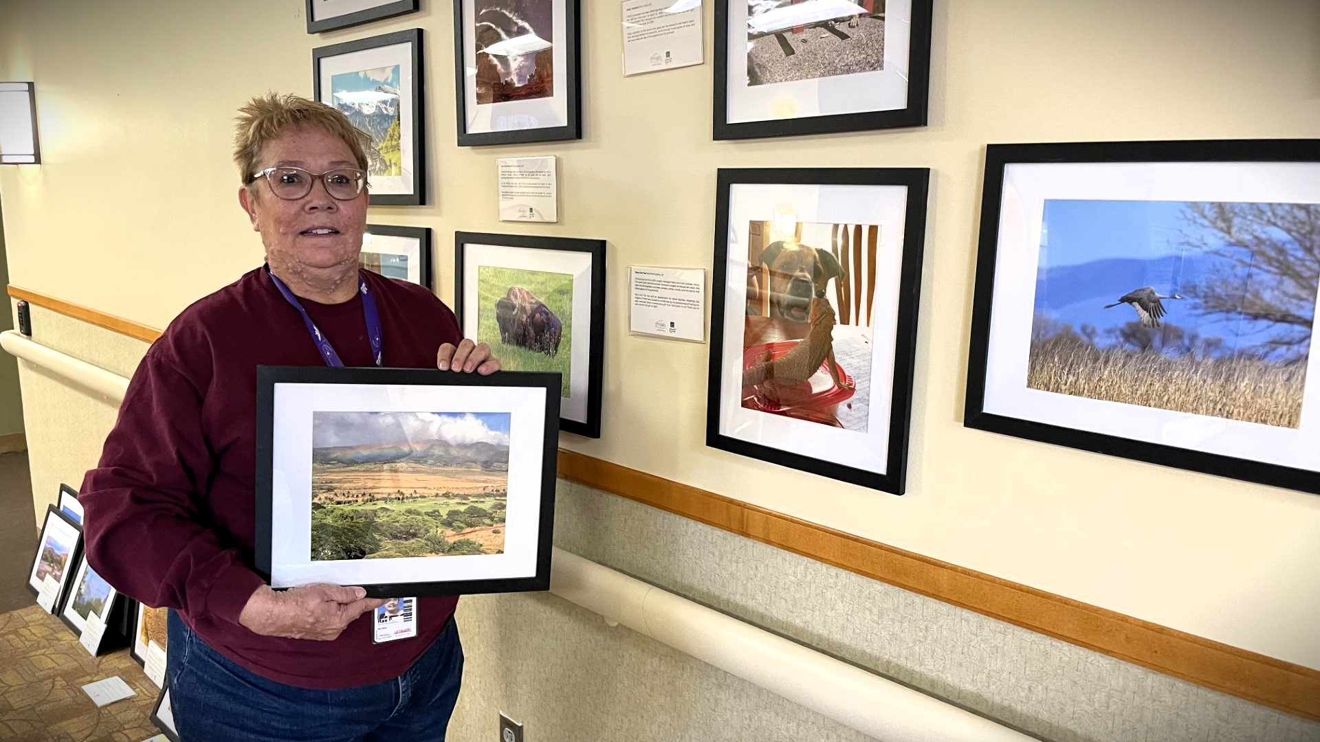 Person holding a framed landscape photo stands beside a wall displaying various framed photographs.