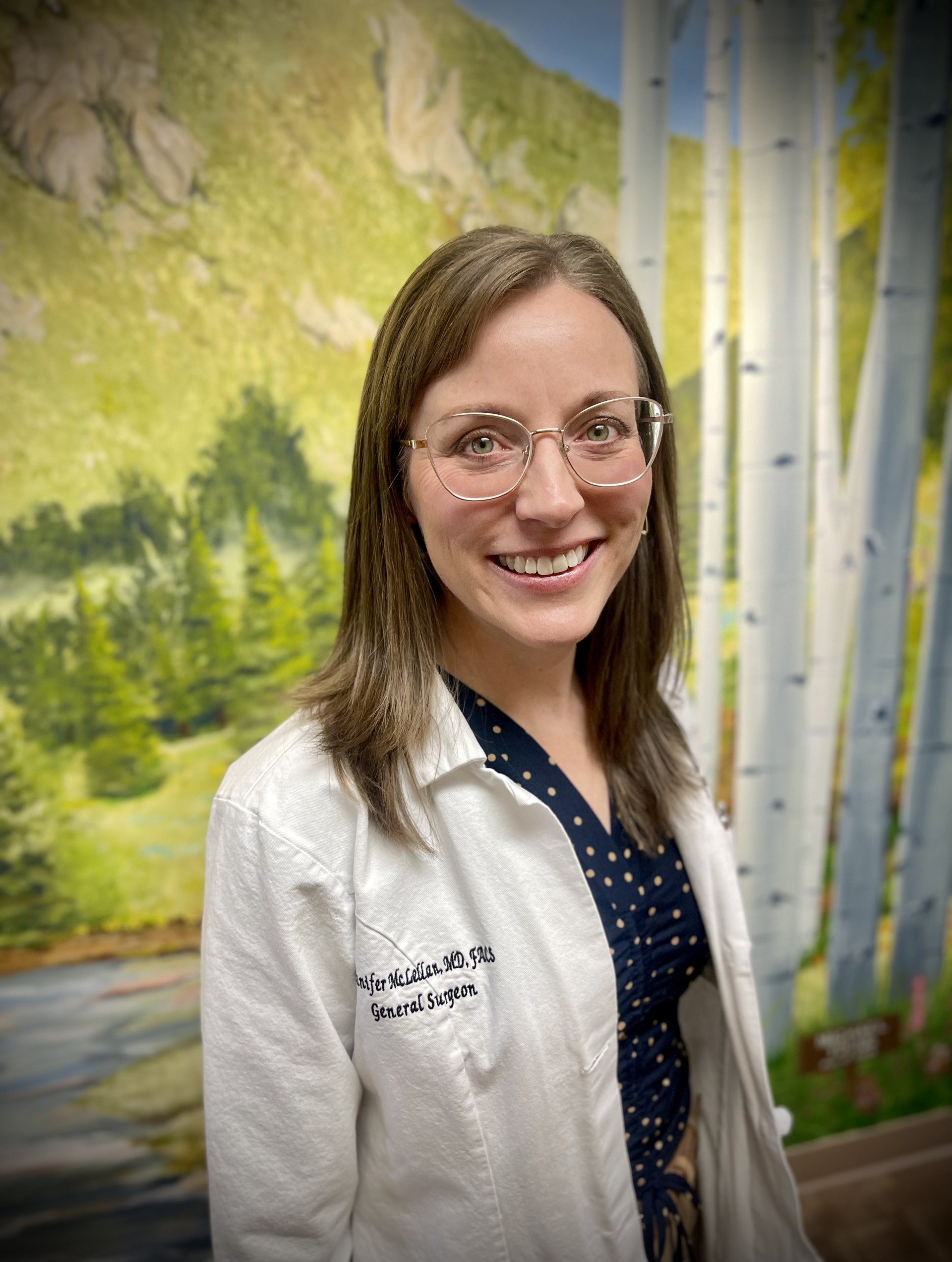 A woman wearing glasses and a white coat stands in front of a mural featuring trees and mountains.