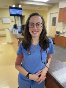 A person in blue scrubs and glasses stands in a hospital setting, smiling, with a stethoscope around their neck.