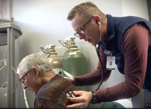 A healthcare professional listens to the back of an elderly man's chest with a stethoscope. Oxygen tanks are visible in the background.