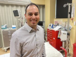 A man in a plaid shirt smiles in a medical room with equipment, including monitors and a red cart, in the background.