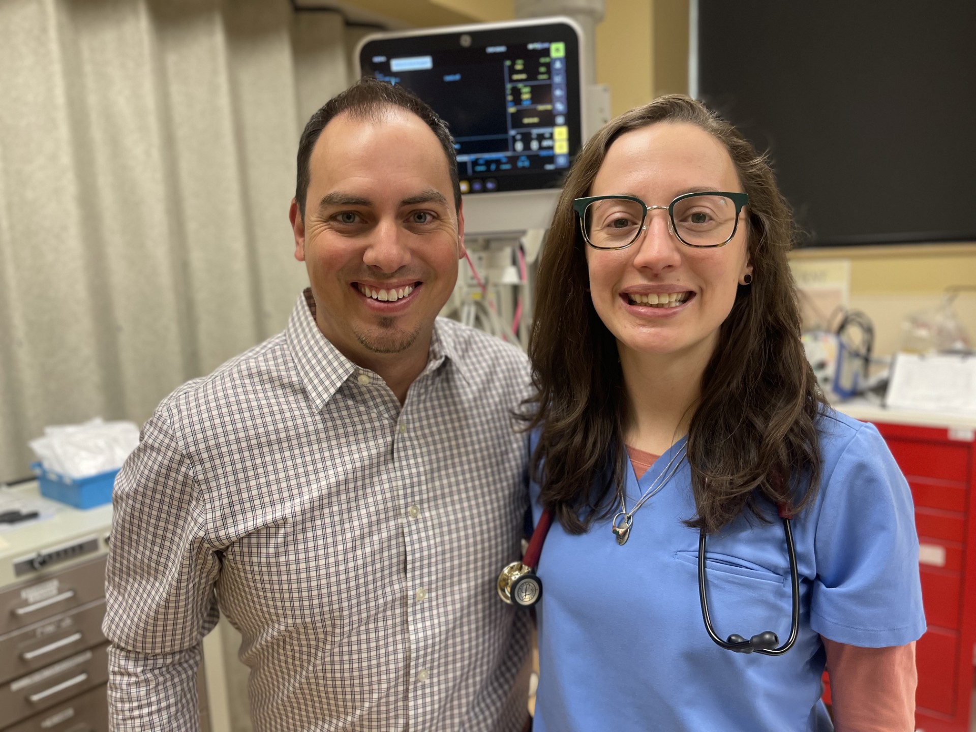 A man in a checked shirt and a woman in blue scrubs with a stethoscope smile while standing together in a medical setting.