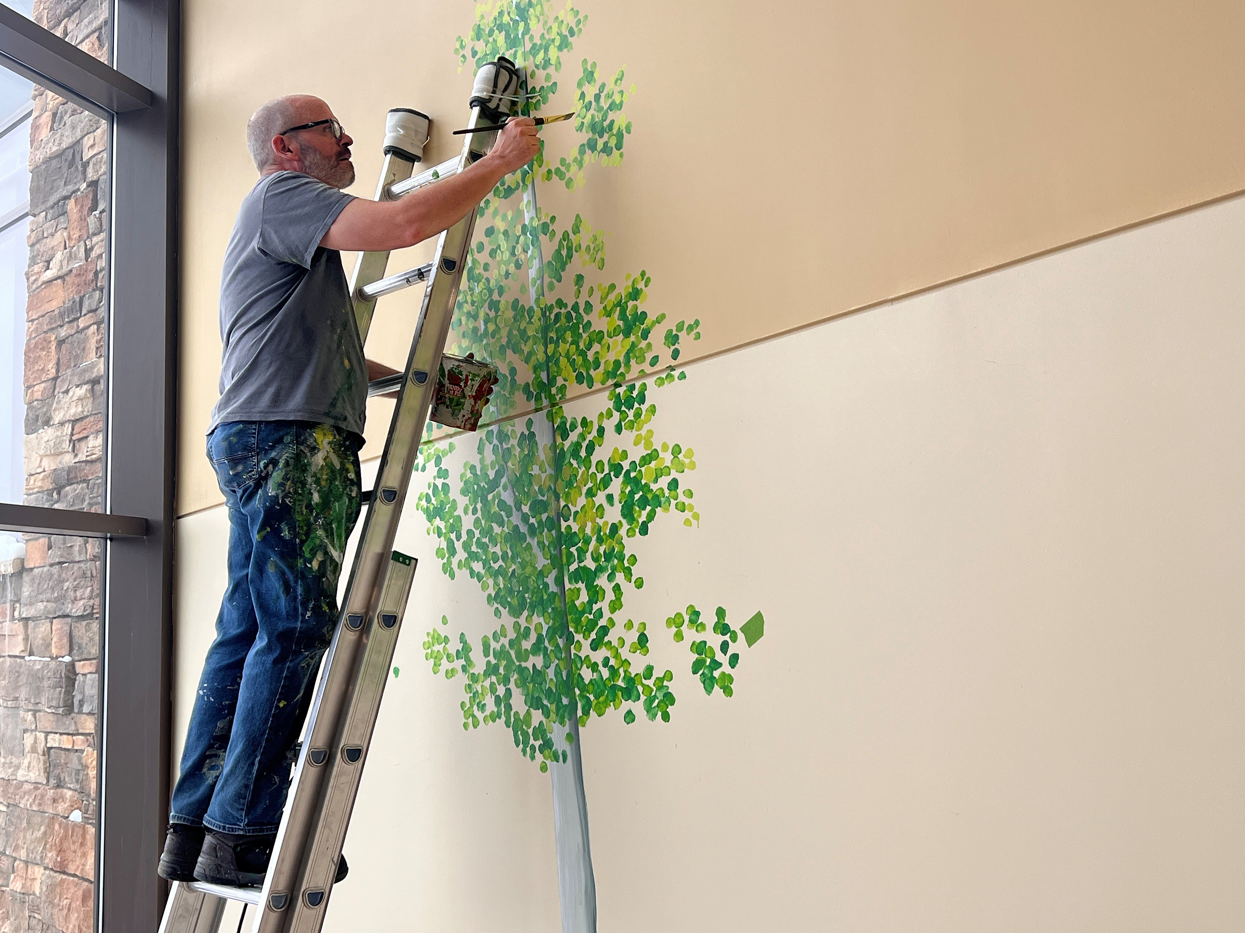 A man standing on a ladder paints a tree mural with green leaves on a beige indoor wall near a large window.