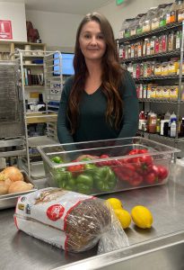 A woman stands in a commercial kitchen behind a container of green and red bell peppers, with onions, lemons, and a loaf of bread on the counter. Shelves with spices are in the background.