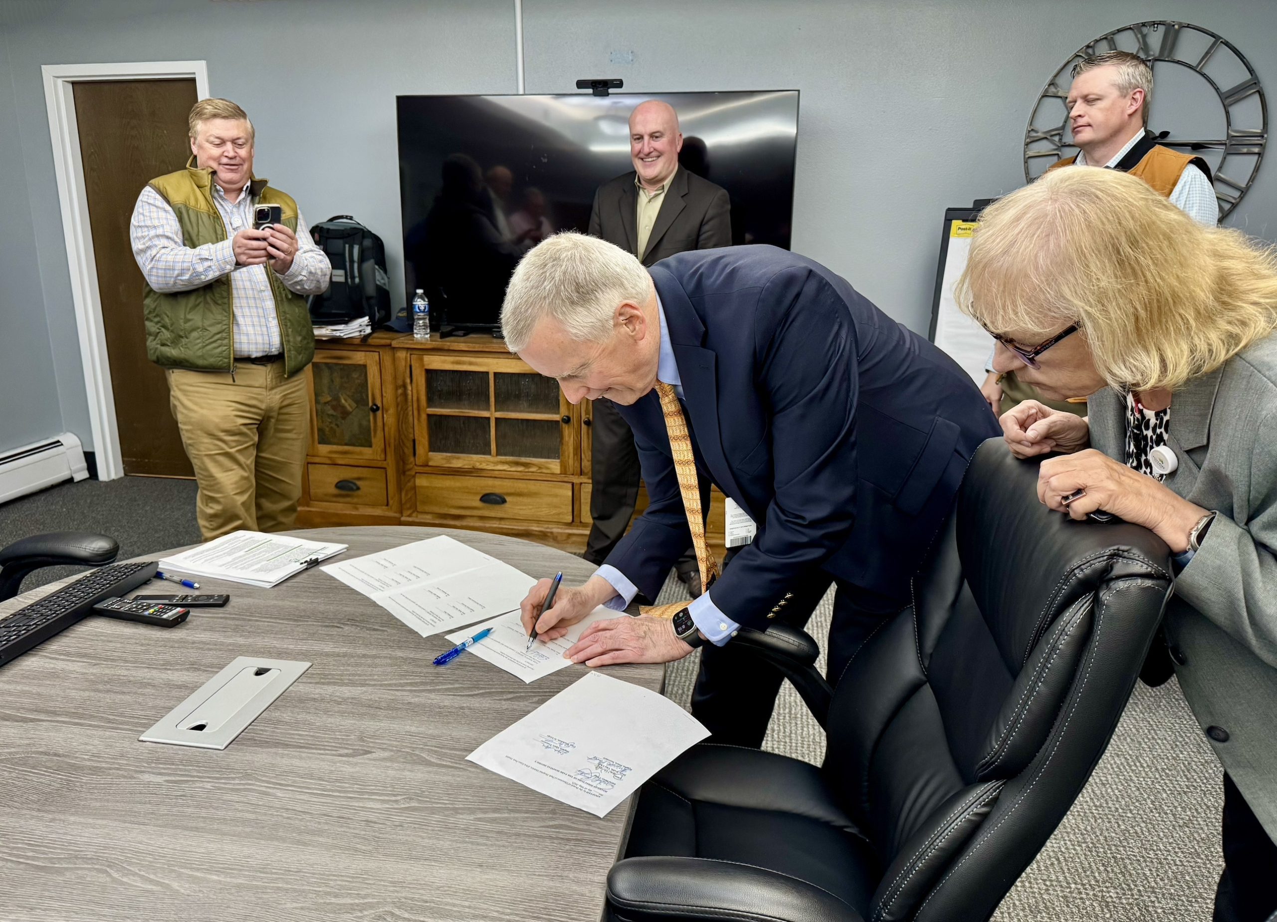 A man in a suit signs documents on a conference table while three people observe and one person takes a photo with a phone.