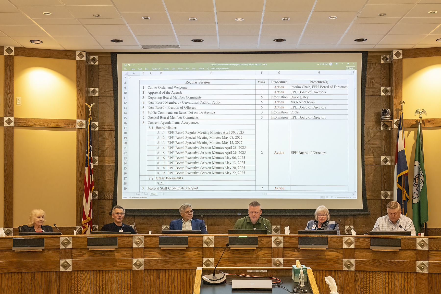 Six people sit at a long desk in a meeting room, with a projected agenda displayed on a screen behind them.