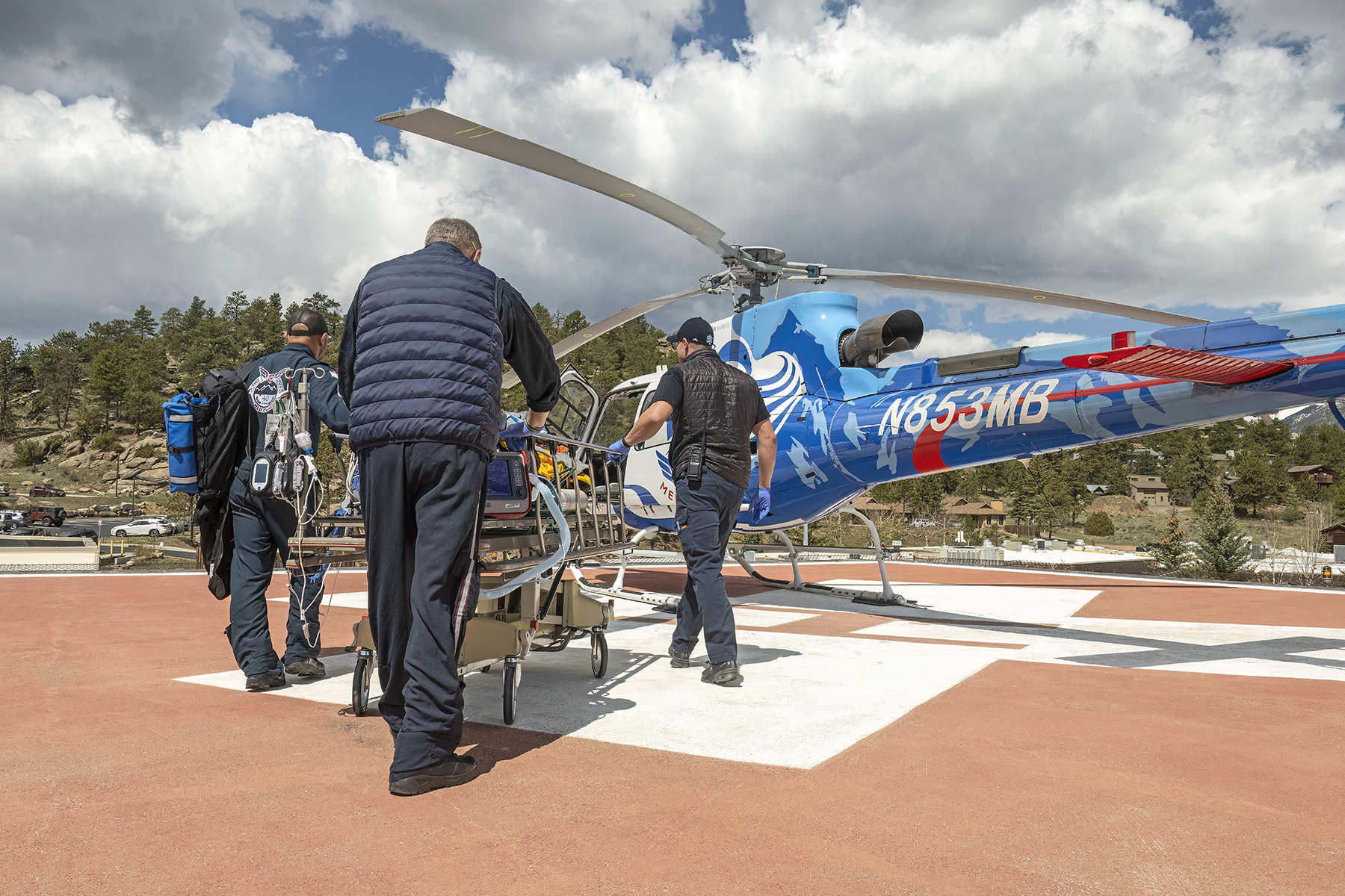 Medical personnel wheel a patient on a stretcher toward a blue helicopter on a helipad under a cloudy sky.