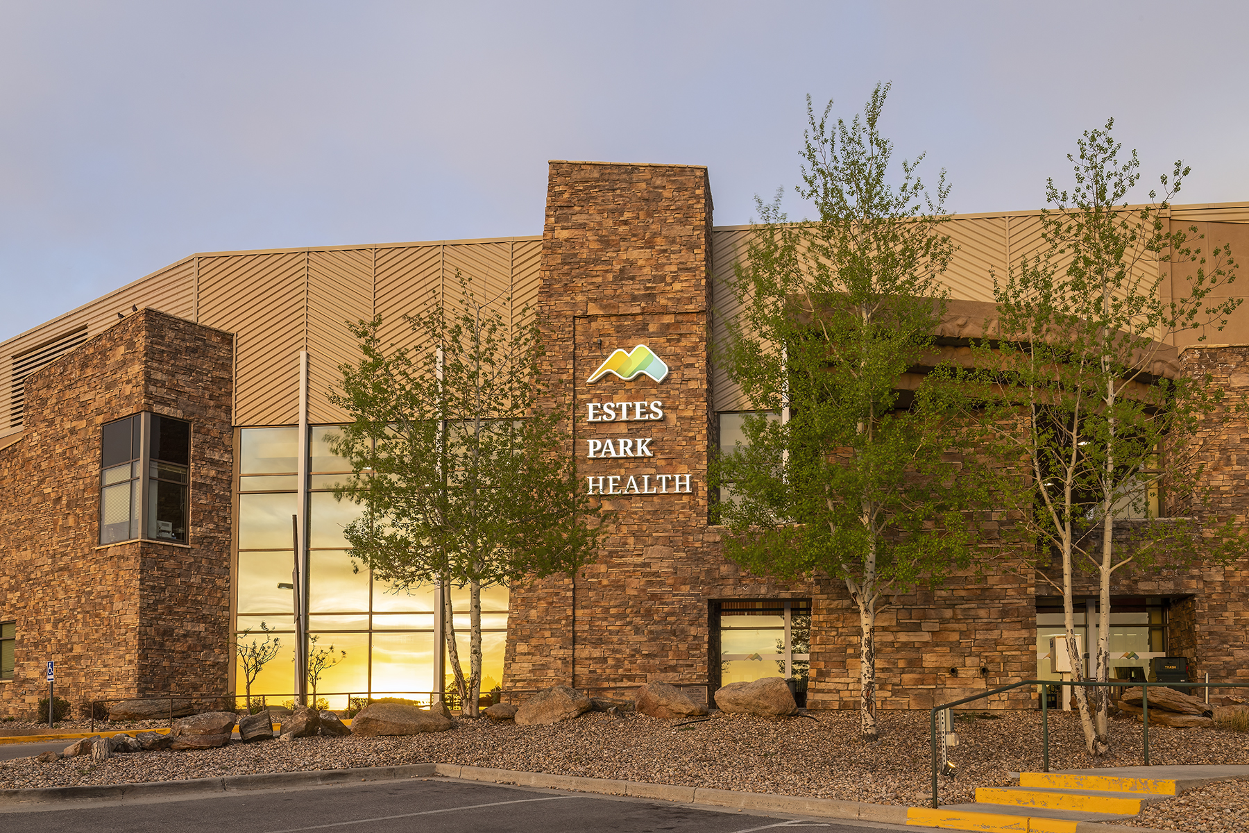 Exterior view of Estes Park Health building with stone facade, large windows, and trees in front, taken at sunset.