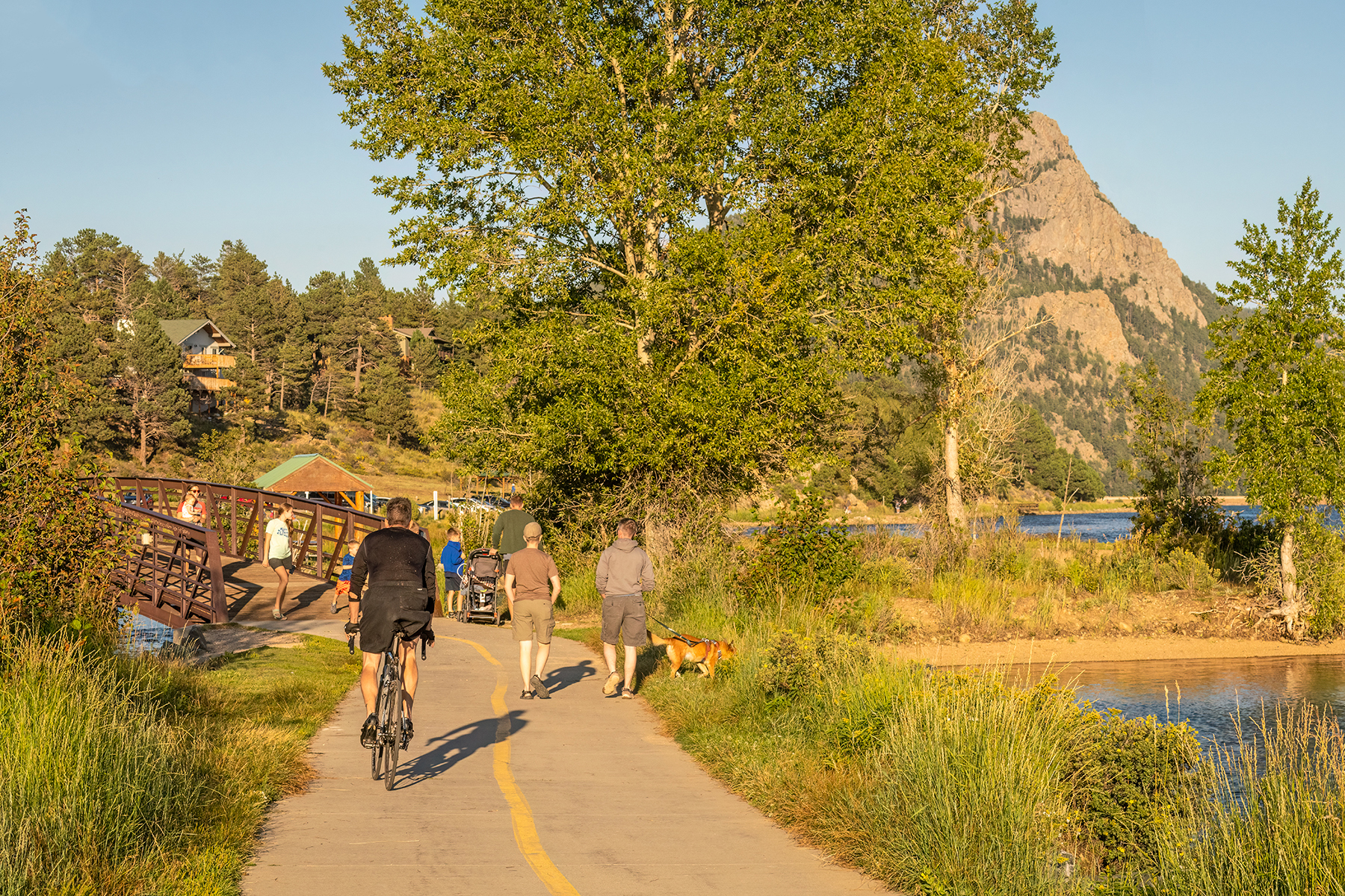 People walk and bike on a paved path near a lake, with trees and a mountain in the background on a sunny day.