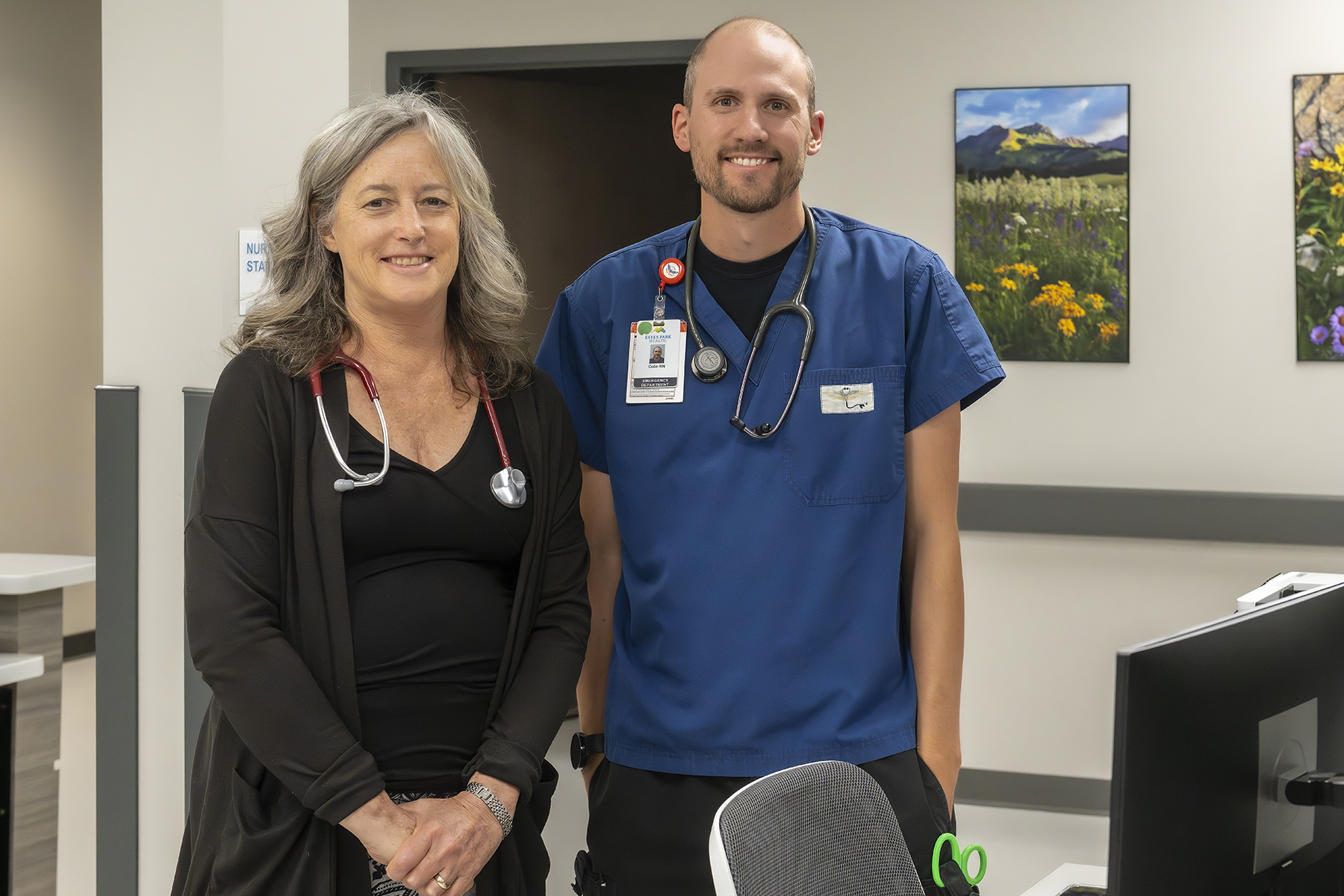 Two healthcare professionals, one in a black outfit and one in blue scrubs, stand in a medical office with medical equipment and nature photos on the wall behind them.