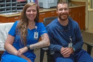 Two healthcare professionals in blue scrubs sit side by side in an office, smiling at the camera. Both are wearing ID badges and seated in front of desks and office equipment.