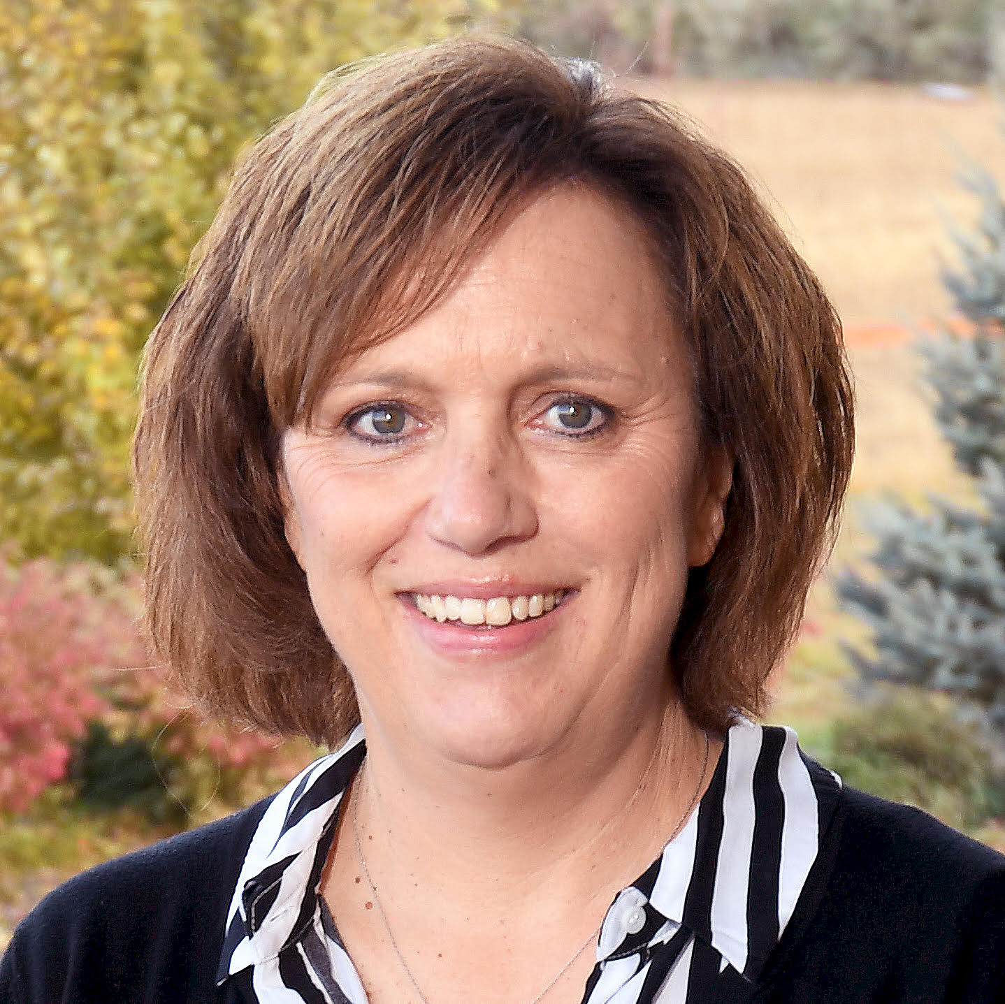A woman with short brown hair and a striped shirt smiles outdoors with trees and greenery in the background.