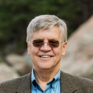 Older man with short gray hair and sunglasses, wearing a blue shirt and brown blazer, smiling outdoors with trees and rocks in the background.